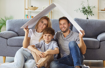 Happy mother, father, and son sit together at home, holding a symbolic roof over heads at home....