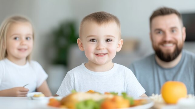 A smiling toddler sits center, in a white shirt, at a table with food. A girl and a man are blurred in the background. The photo is well-lit, high-quality, and features warm tones. Its a candid, happ