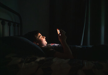 Teen boy in bedroom using smartphone while laying on his bed at night.