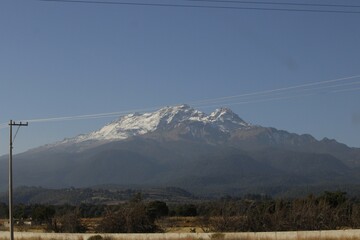 paisaje de monta&ntilde;a con nieve, arboles y pastizales, con poste de luz en la carretera y fondo de cielo azul