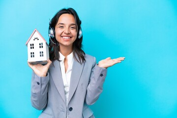 Real estate hispanic agent holding a toy house isolated on blue background extending hands to the...