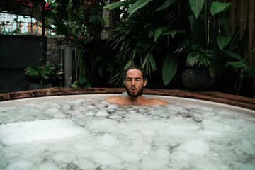 Man takes an ice bath outdoors in a lush tropical setting. Cold immersion for mental strength,...