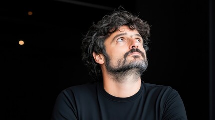 Obraz premium A high-resolution portrait shows a man with dark, curly hair and a beard, looking upward. He wears a simple black t-shirt. The background is dark, providing a stark contrast. The image is well-lit,