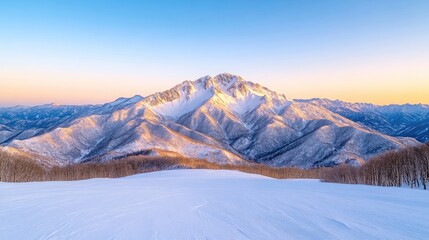 A snow-covered mountain range dominates this landscape photograph. The main peak is sharply defined against a clear, sunset sky. The foreground shows a smooth, snow-covered slope. The image is high-