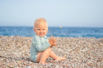 A joyful and cheerful toddler joyfully sits on a pebbly beach, smiling brightly with the vast ocean in the background