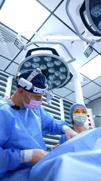 Caucasian surgeon uses a tool device shoving it back and forth quickly. Scrub nurse in yellow glasses at backdrop. Low angle view. Vertical video.