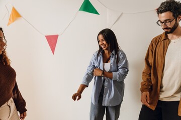 A young Black woman in her late 20s, rolling up her sleeve while laughing with her friends as she stands against a white wall with triangle cloth decorations on it