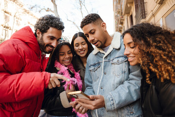 Five friends in their 20s stand close together outdoors on a city street, looking at a smartphone held by a Hispanic man in a denim jacket, with buildings in the background, on a sunny day.