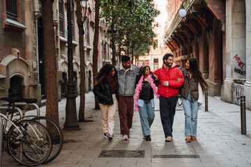 Obraz premium A multiethnic group of friends in their late 20s dressed in jackets walking in a row on a cozy street with old buildings and three on the sides, a woman in the middle is in a crown and a feather scarf