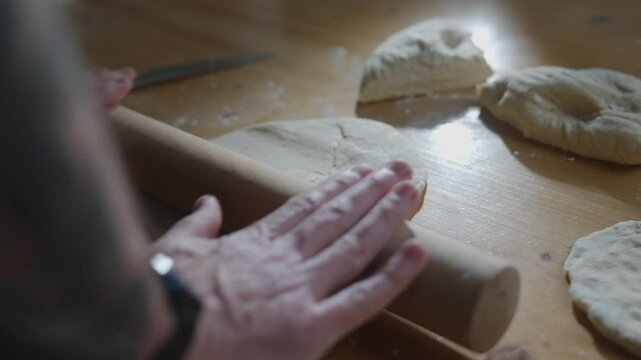 A person is making a pizza dough on a wooden table. The dough is being rolled out with a rolling pin