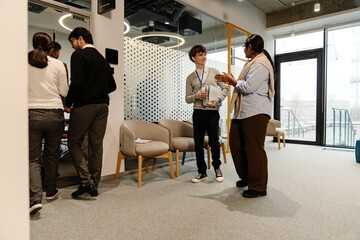 In an office, a multiethnic group of formally dressed people is entering a meeting room, with a White man in his 20s, holding documents and talking to an Indian woman in her late 20s who gestures.