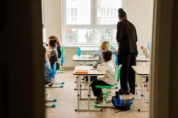 A young, school-aged White boy, sitting at his desk in a busy classroom, turning back to look at one of his classmates, as his mid-30s Black female teacher stands beside him © Drobot Dean
