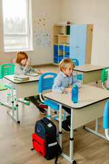 Obraz premium Two elementary school White students sit at desks and work on their studies in a classroom. The room contains colorful furniture, shelves, decorations, backpacks, and school supplies.