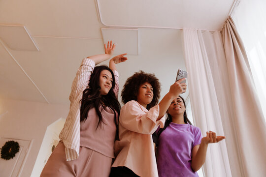 A low angle of three multiethnic women in their 20s, one Asian, Black with an afro, and another Black smile and pose in pajamas while taking a selfie together using a smartphone, at home near curtains