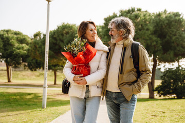A smiling casually dressed White couple in their 50s, a brunette woman holding a wrapped olive tree seedling, and a man with graying hair and a beard, walk together and talk in a park, on a sunny day.