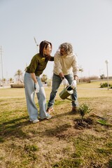 A middle-aged White man in his late 50s, watering a small plant as his mid-50s White wife encourages him while holding him by the shoulder, as they stand together outside on a grassy plain