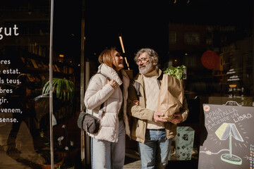 A White wife in her late 50s, talking to her middle-aged White husband who holds a grocery bag, as they're walking out of a book store, on a sunny day, on their date