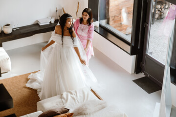Two young women, a Latino in a white wedding dress and a Caucasian in a pink floral dress, stand in a room with large windows. They are trying to get the best dress for the upcoming wedding.