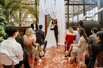 A wholesome Black bride and groom in their late 20s, kissing as they stand in front of their wedding guests, finishing the ceremony of their wedding, in a large chamber with potted plants all around
