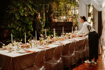 Two young White women, one in a black jacket and the other in a white shirt, arrange a formal dining table. The table is set with candles, flowers, and various dishes near lush greenery.