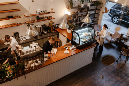 An overweight White woman in her 20s, standing behind the counter at a cozy coffee shop and writing something in her notepad, as her two adult White colleagues, a man, and a woman, work around her