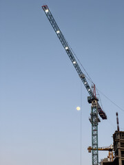 Naklejka premium dusk construction site, a tall construction crane is lit up against a clear sky at either dawn or dusk, with the moon adding to the scene a building under construction is visible in the background