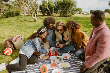 A young Asian woman in her late 20s, showing something on her phone to a group of her young diverse friends, as they sit together on a blanket on a grassy field, having a picnic, on a sunny day