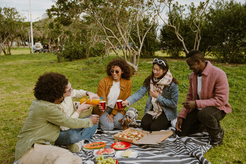 A diverse group of five young friends, all in their 20s, having a picnic together on a grassy field in the park, pouring some soda into their plastic cups and chatting, with pizza on the blanket