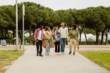 Five multiethnic friends walk together through a park. They wear colorful casual outfits, and an adult Black man with an afro and green shirt carries a camera. They converse and enjoy their walk.
