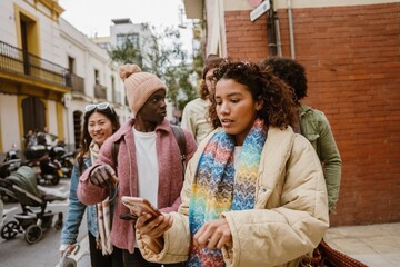 A young Latina woman in her late 20s, looking at her phone with a shocked expression on her face, as she stands on a city street alongside a diverse group of her young friends, on a sunny day