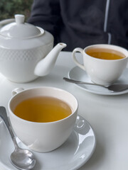 tea time scene, on a table is a teapot and two filled cups, one with a saucer and spoon in the foreground and a person in a dark jacket in the background