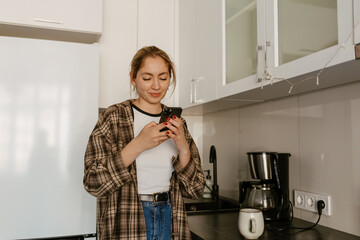 A young White woman in her late 20s, standing in a modern kitchen and looking at her phone with a...