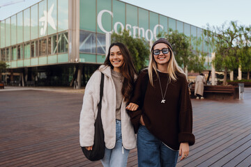 In an urban plaza with a wooden deck before a modern glass building, two smiling women in their 20s, one Asian brunette in a white jacket, and a White blonde in a brown sweater, walk arm-in-arm.