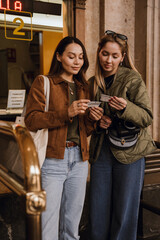Two casually dressed smiling women in their late 20s, a White blonde and an Asian brunette, stand...