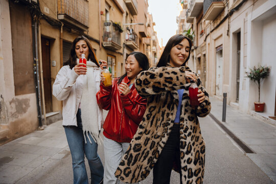 Three diverse women walk down a city street enjoying drinks and each other's company. One wears a white coat, another a red jacket, and the third a leopard-print coat during their conversation.