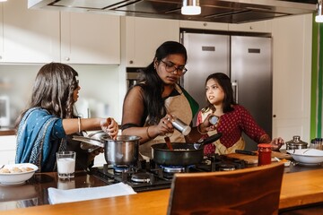 An Indian woman in her late 30s, salting the water in the pot as she cooks in a modern kitchen together with her two younger sisters, with the light hitting them from the side