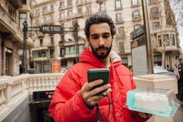 An adult White man in a red puffer jacket stands at a metro station entrance. He holds a smartphone...