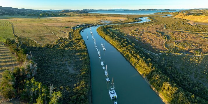 Aerial view of sailboats moored along a river in a rural landscape. The boats are lined up, reflecting in the calm water. Taken in New Zealand. CLEVEDON, NZ