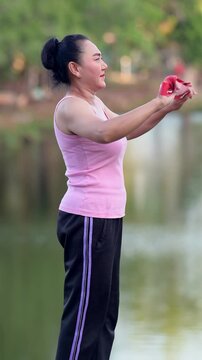 Middle Aged Asian Woman In Side Profile Exercises Outdoors With Resistance Band, Sporting Bun And Athletic Tank Top Near Water, Focusing On Body Fitness