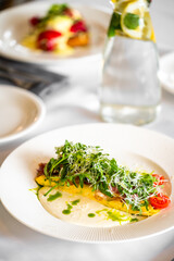 A beautifully plated dish featuring a vibrant salad with fresh greens, cherry tomatoes, and drizzled sauce. In the background, a bottle of water with lemon and mint adds a refreshing touch.