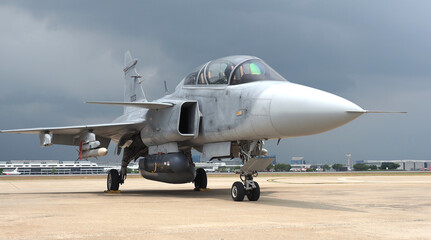 A military fighter jet parked on an airstrip under cloudy skies. The aircraft features a sleek design