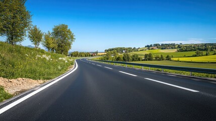 Fototapeta premium Scenic asphalt road curves through a vibrant green countryside under a clear blue sky.