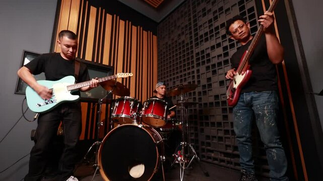 Three Asian male musicians playing electric guitar, bass and drums in soundproof music studio, focusing on live performance and teamwork in modern indoor setting