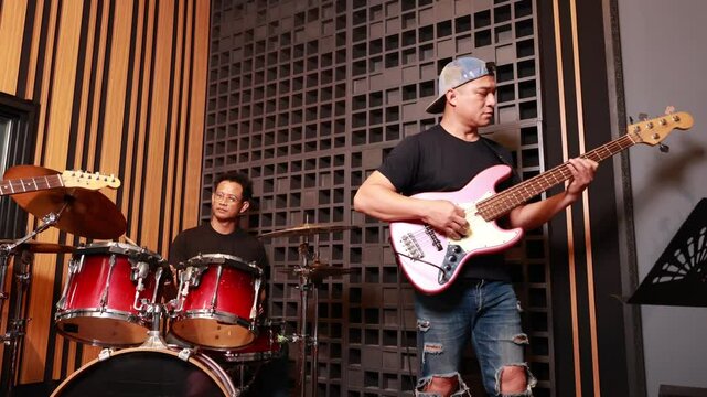 Asian male musicians smiling and engaging while playing bass, drums and guitar in soundproof recording studio, showing teamwork and enjoyment during rehearsal