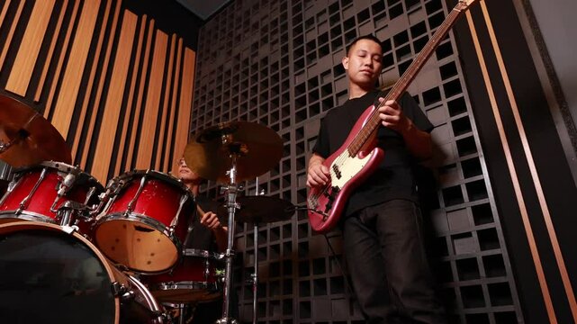 Two young Asian men playing drums and electric bass in professional recording studio, focused on performing music, warm light, black t-shirts, modern soundproof wall