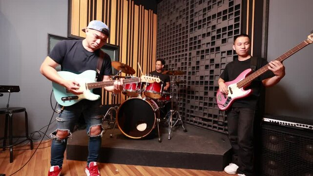 three asian men performing together in professional soundproof studio, electric guitarist in foreground, drummer and bass guitarist in background, focused and energetic rehearsal session