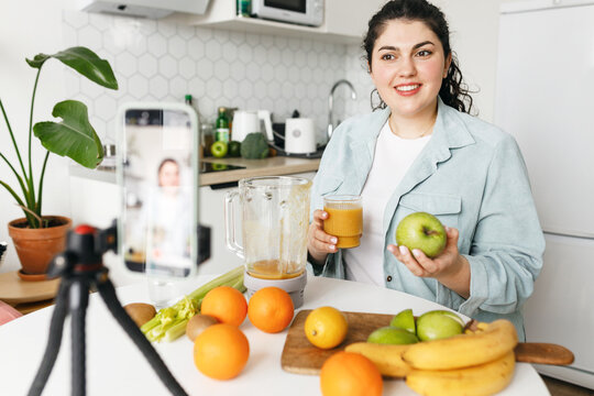 Selective focus on young plus size woman holding apple in one hand and glass of fresh smoothie in another recording video on smartphone or having live on social media, sharing recipe for weight loss
