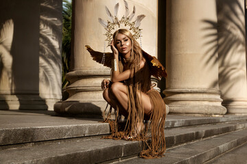 Beautiful sexy blonde model in luxury macrame dress, gold wings and goddess headband poses...