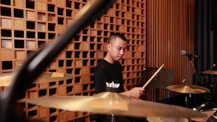 focused young asian male drummer playing drum set in acoustic recording studio, hitting cymbals with drumsticks, indoor performance setup with microphones and wooden panels