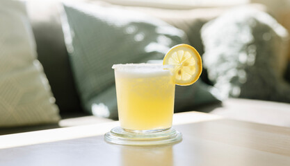 Classic summer drink Margarita cocktail with salt rim and lemon wheel on wooden table in living room under sunlight through the window.